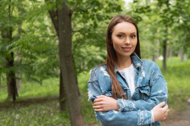pretty young student in denim jacket looking at camera while standing with crossed arms in park clipart
