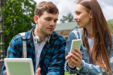 Dijital tabletli yakışıklı öğrenciye akıllı telefon gösteren neşeli kız. 