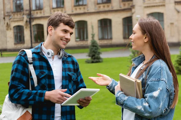 Radosna Dziewczyna Gestykulująca Trzymając Książki Rozmawiając Szczęśliwym Studentem Trzymającym Tablet — Zdjęcie stockowe