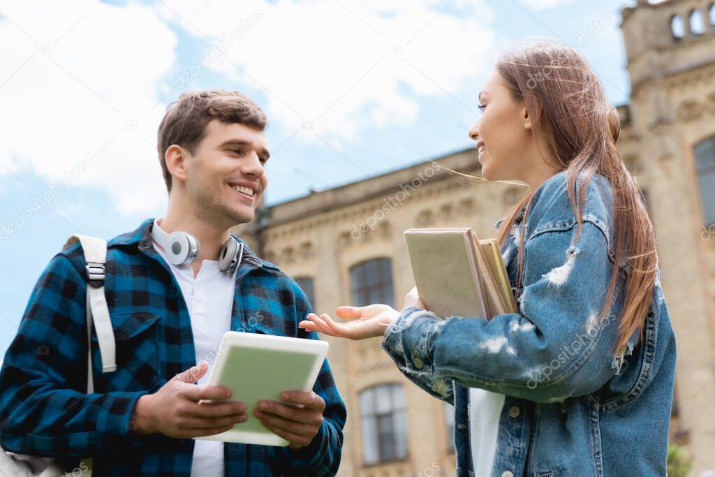low angle view of cheerful girl gesturing while holding books and talking with happy student 