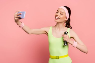 Neşeli sporcu kadın dambılı tutuyor ve pembe selfie çekiyor. 