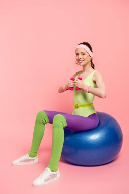 positive sportswoman exercising with dumbbells and sitting on fitness ball on pink  clipart