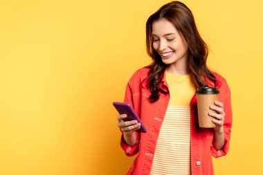 Mutlu genç bir kadın elinde kahve tutarken sarıda akıllı telefon kullanıyor.