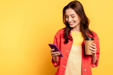 happy young woman holding coffee to go and using smartphone on yellow clipart