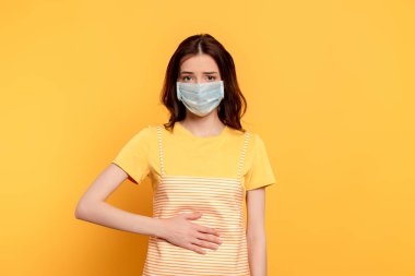 sick young woman in medical mask having stomach ache on yellow clipart