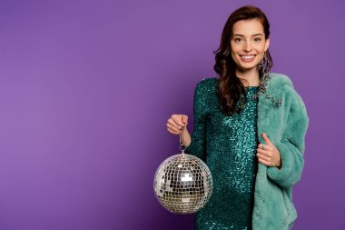 cheerful and stylish woman holding disco ball and showing thumb up on purple  clipart