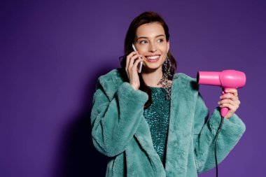 cheerful girl talking on smartphone of holding hair dryer on purple  clipart
