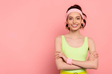 Saç bandı takmış mutlu genç bir kadın kollarını kavuşturup pembe üzerinde gülümsüyor. 