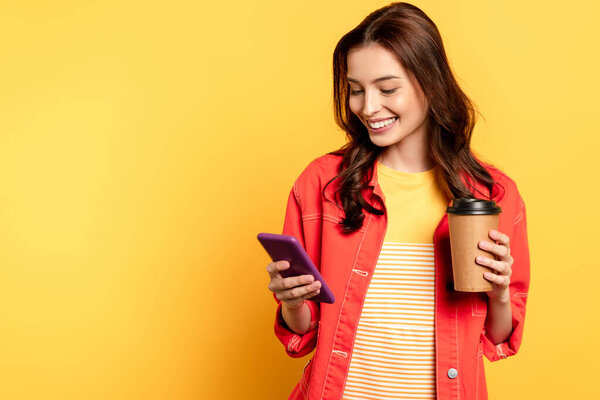 happy young woman holding coffee to go and using smartphone on yellow