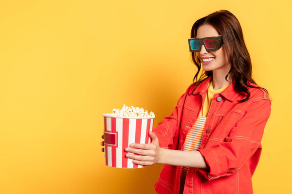 happy young woman in 3d glasses looking at popcorn bucket on yellow