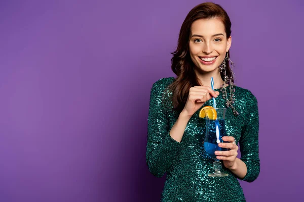 cheerful woman in party dress holding alcohol cocktail and straw on purple