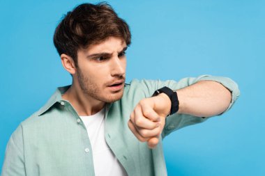 Şok olmuş genç adam mavi üzerine izole edilmiş kol saatine bakıyor.