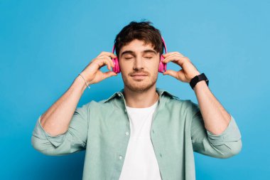 Mutlu genç adam mavi kanalda müzik dinlerken kablosuz kulaklıklara dokunuyor.