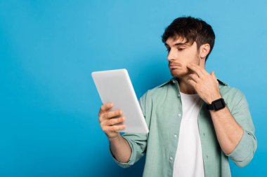 Düşünceli genç adam mavi tablet üzerindeki dijital tablete bakarken yüze dokunuyor.