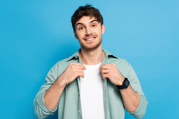 handsome young man touching shirt while smiling at camera on blue