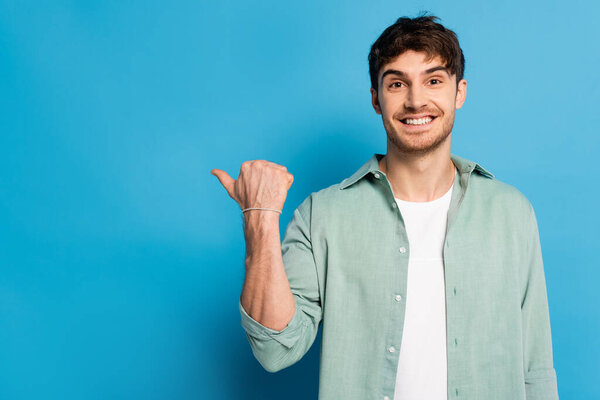 happy young man pointing with thumb while looking at camera on blue