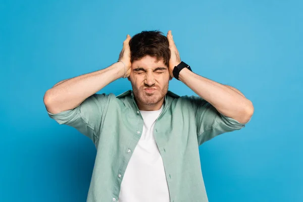 stressed young man touching head while suffering from headache on blue