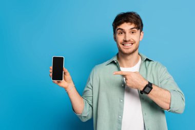 Mutlu genç adam mavi ekranlı akıllı telefonu işaret ediyor.