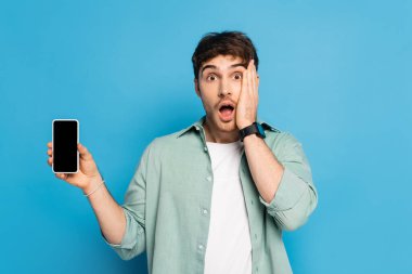 Şok olmuş genç adam yüzüne dokunuyor ve mavi ekranda boş ekranlı akıllı telefonu gösteriyor.
