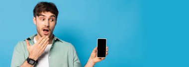 Şok olmuş genç adamın mavi ekranda boş ekranlı akıllı telefonu gösteren yatay görüntüsü