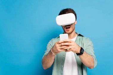 Şok olmuş genç adam mavi üzerine akıllı telefon kullanıyor.