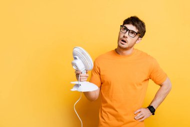 displeased young man looking up while suffering from heat and holding electric fan on yellow clipart