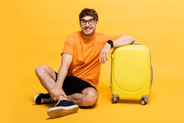 Sarı yaz tatili için seyahat çantasının yanında oturan neşeli erkek turist. 