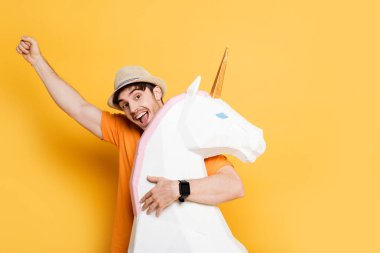excited man in summer hat holding fist up and hugging decorative unicorn on yellow  clipart