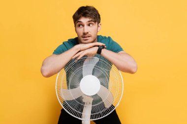 Düşünceli genç adam elinde elektrikli vantilatörle duruyor.     