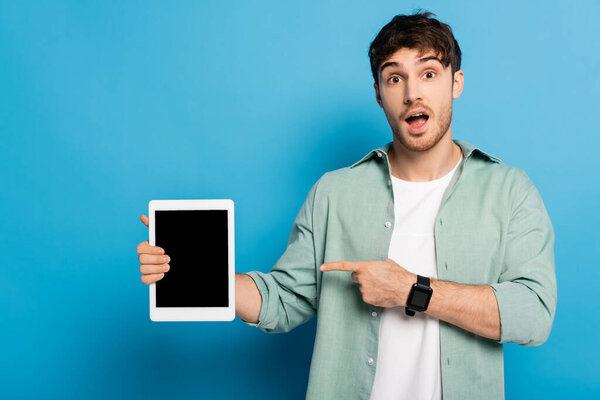 shocked young man pointing at digital tablet with blank screen on blue