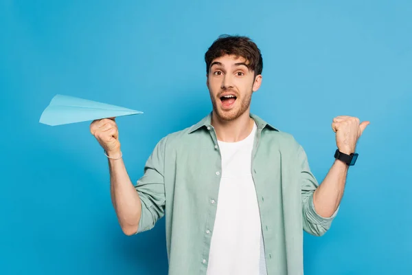 Excited Young Man Pointing Thumb While Holding Paper Plane Blue — Stock Photo, Image