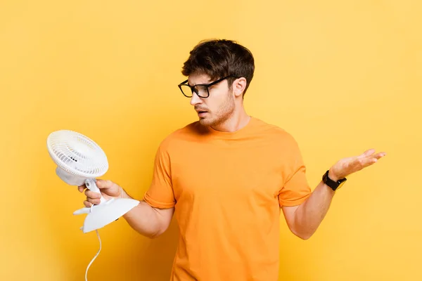 Förvirrad Ung Man Visar Ryckig Gest Medan Håller Elektrisk Fläkt — Stockfoto