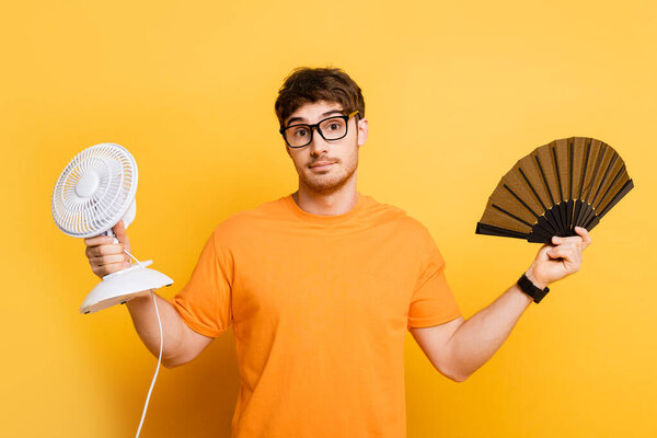 discouraged young man holding electric and hand fans while looking at camera on yellow