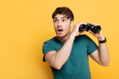 handsome shocked man holding binoculars on yellow clipart