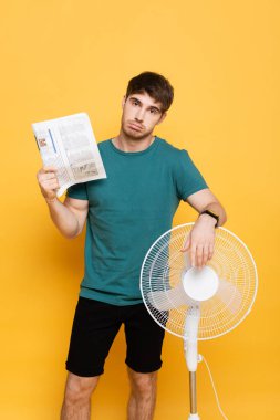 Üzgün adam elektrikli yelpaze ve gazeteyle sıcaktan muzdarip. Sarı yelpaze gibi.    