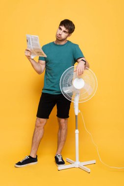 Kafası karışmış bir adam elektrikli yelpaze ve gazeteden muzdarip sarı yelpaze gibi    