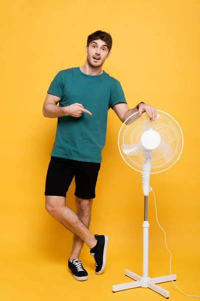 Jovem Surpreso Apontando Com Dedo Para Ventilador Elétrico Amarelo — Fotografia de Stock