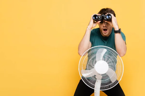 Homem Chocado Olhando Através Binóculos Enquanto Com Ventilador Elétrico Amarelo — Fotografia de Stock