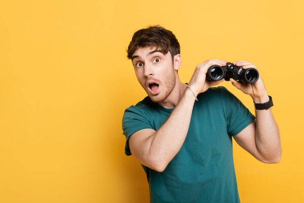 handsome surprised man holding binoculars and looking at camera on yellow