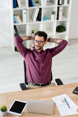 Gülümseyen iş adamının yüksek açılı görüntüsü elleri başlarının arkasında boş ekranlı, dizüstü bilgisayarlı ve kağıtlı dijital tabletin yanında oturuyor.