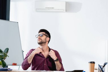 İş yerinde oturan yorgun işadamı ofiste sıcaktan acı çekerken gömleğe dokunuyor.