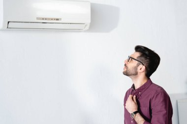 Genç işadamı gömleğe dokunurken ve sıcaktan acı çekerken klimaya bakıyor.
