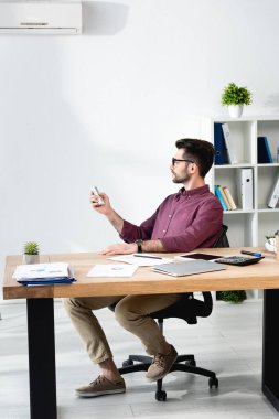 Yakışıklı iş adamı işyerinde oturuyor ve klimayı uzaktan kumandayla açıyor.
