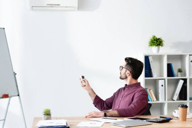 Genç işadamı işyerinde oturuyor ve klimayı uzaktan kumandayla açıyor.