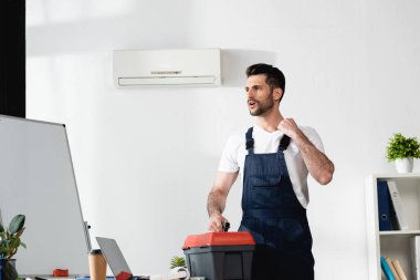 handsome repairman standing with toolbox near broken air conditioner and suffering from heat clipart