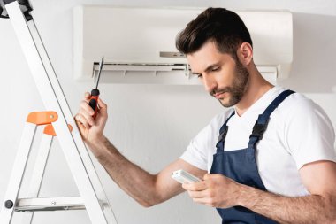 Uzaktan kumanda ve tornavida tutan yakışıklı bir tamirci klimanın yanında merdiven başında dikilirken