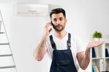 Akıllı telefondan konuşan kafası karışmış bir tamirci ve duvardaki klimanın yanında omuz silkme hareketi gösteriyor.