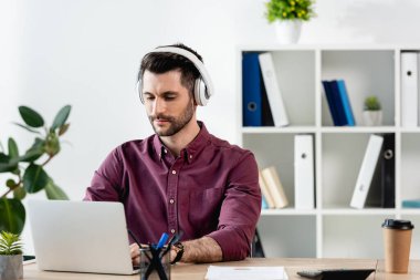 İşyerinde laptopta çalışan kablosuz kulaklıklı dikkatli iş adamı.