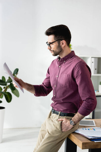 young, serious businessman reading document while leaning on desk with hand in pocket