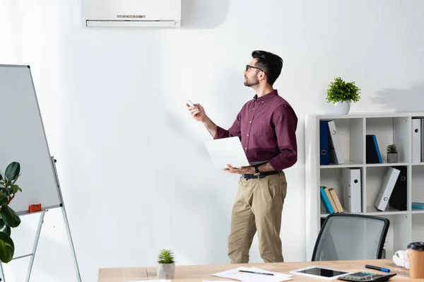 Young Businessman Holding Laptop Switching Air Conditioner Remote Controller — Stock Photo, Image